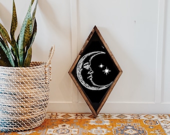 Op de afbeelding: Een zwart, ruitvormig houten frame met een wit halve maan- en sterrenontwerp op een zwarte achtergrond. Het frame leunt tegen een witte muur en op de voorgrond staat een geweven mand met een groene plant.