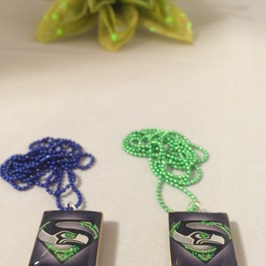 May include: Two rectangular pendant necklaces with a team logo, one with a blue beaded chain and the other with a green beaded chain. The pendants have a dark blue background with a team logo in silver, green, and white.