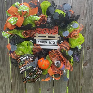 Op de afbeelding: Een feestelijke Halloween-krans met oranje, groene en zwarte mesh, geaccentueerd met oranje en groene stippenlinten. Voorzien van een bord met "PUMPKIN PATCH" en een kleine oranje pompoen decoratie.