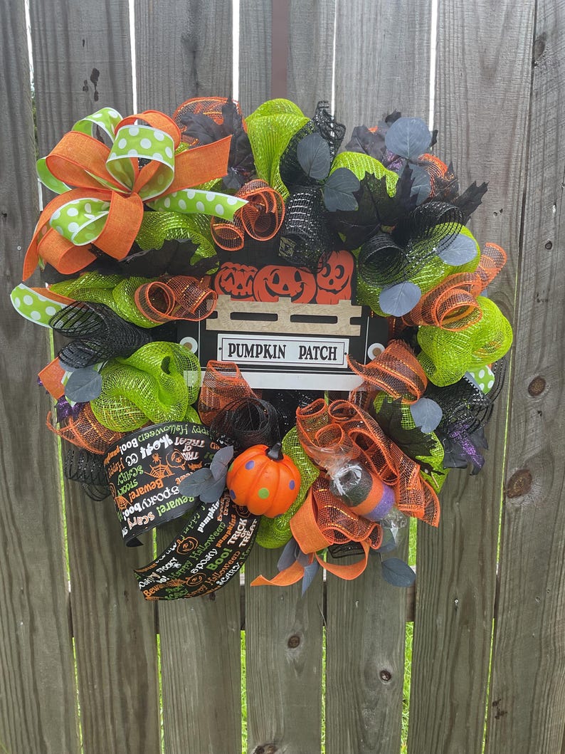 Op de afbeelding: Een feestelijke Halloween-krans met oranje, groene en zwarte mesh, geaccentueerd met oranje en groene stippenlinten. Voorzien van een bord met "PUMPKIN PATCH" en een kleine oranje pompoen decoratie.