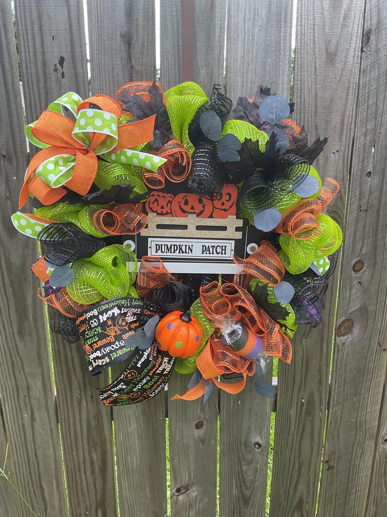 Op de afbeelding: Een Halloween-krans met oranje, groene en zwarte mesh, linten en decoratieve elementen. Voorzien van een bord met de tekst "PUMPKIN PATCH" en een kleine pompoen. Bevat tekst met de tekst "Beware! Scary" en "Happy Halloween Boo!"