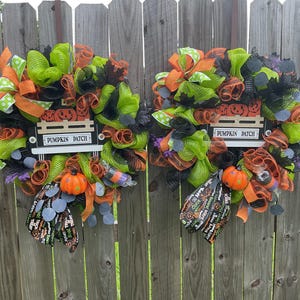 Op de afbeelding: Twee Halloween-kransen met groene, oranje en zwarte mesh, linten en decoratieve elementen. Elke krans heeft een bord met de tekst "Pumpkin Patch", een kleine oranje pompoen en een decoratieve tas. De kransen hangen aan een houten hek.