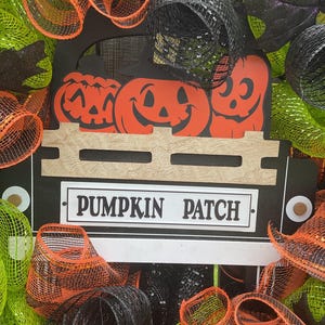 Op de afbeelding: Een Halloween-krans met een zwart vrachtwagenbedontwerp met oranje pompoenen en een spookuitsparing. Het bord luidt "PUMPKIN PATCH". De krans is versierd met oranje, zwart en groen gaas.