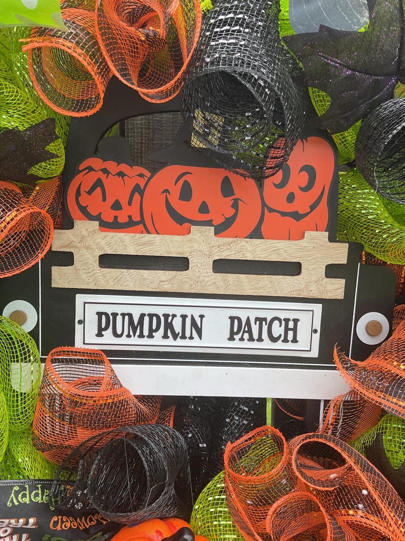 Op de afbeelding: Een Halloween-krans met een zwart vrachtwagenbedontwerp met oranje pompoenen en een spookuitsparing. Het bord luidt "PUMPKIN PATCH". De krans is versierd met oranje, zwart en groen gaas.
