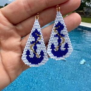 May include: Handmade beaded earrings with a nautical theme. Each earring features a white and blue triangle design with a navy blue anchor and gold-coloured accents. The earrings are suspended from gold-coloured hooks.