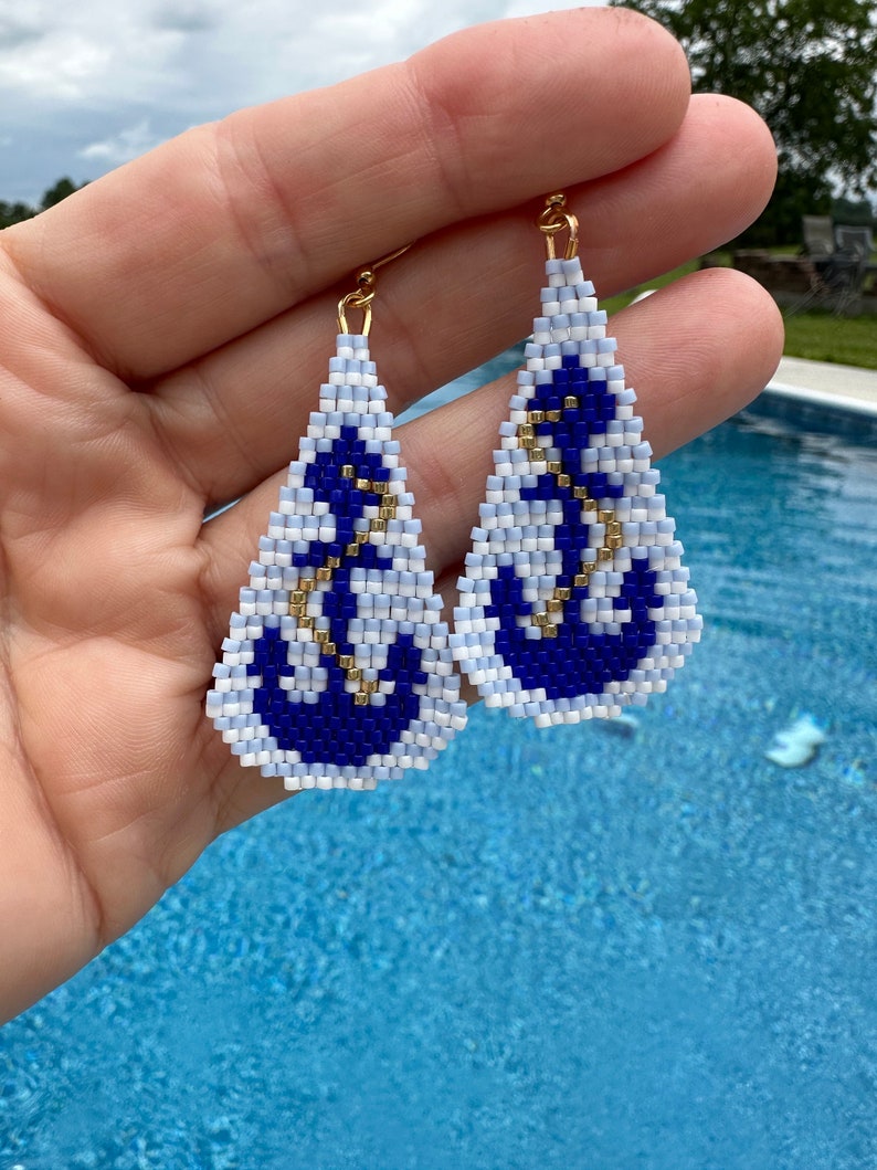 May include: Handmade beaded earrings with a nautical theme. Each earring features a white and blue triangle design with a navy blue anchor and gold-coloured accents. The earrings are suspended from gold-coloured hooks.