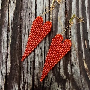 May include: A pair of red beaded heart earrings. The earrings are made of small, rectangular beads and hang from gold-colored posts and chains. The heart shape is elongated and tapers to a point.