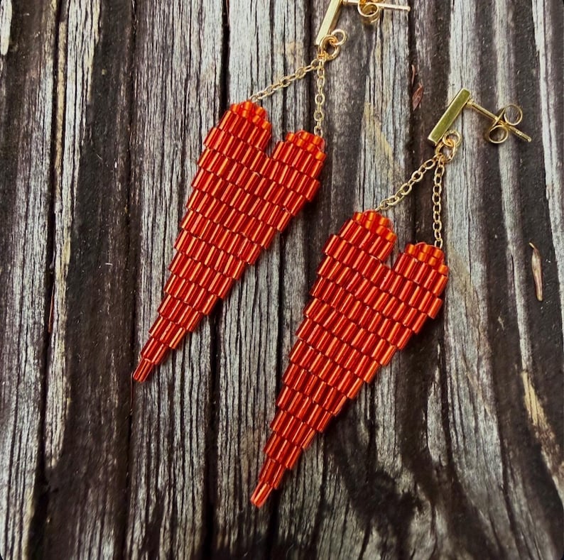 May include: A pair of red beaded heart earrings. The earrings are made of small, rectangular beads and hang from gold-colored posts and chains. The heart shape is elongated and tapers to a point.