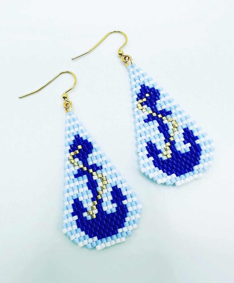 May include: A pair of beaded earrings with a nautical theme. The earrings are teardrop-shaped, featuring a blue anchor design on a light blue and white background. Gold-coloured hooks complete the design.