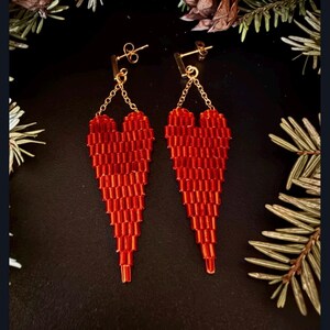 May include: A pair of red beaded heart-shaped earrings. The earrings are suspended from gold-colored posts and chains. The heart design is made of small, rectangular beads, creating a textured appearance. The background is dark, with hints of greenery.