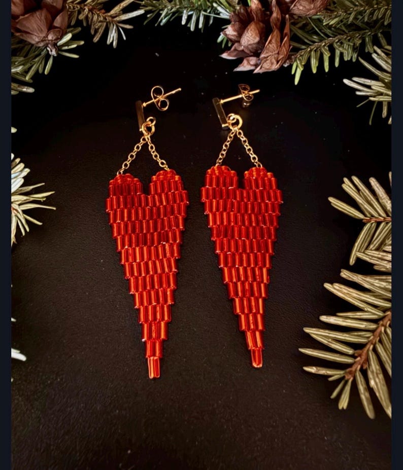 May include: A pair of red beaded heart-shaped earrings. The earrings are suspended from gold-colored posts and chains. The heart design is made of small, rectangular beads, creating a textured appearance. The background is dark, with hints of greenery.