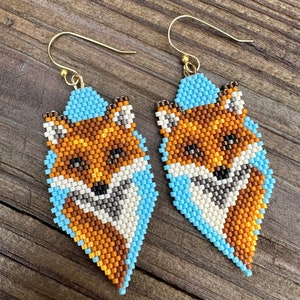 Puede incluir: Un par de pendientes de zorro con cuentas. Los pendientes presentan un dise&ntilde;o de zorro con cuentas naranjas, marrones, blancas y negras. El zorro est&aacute; delineado con cuentas de color azul claro y los pendientes tienen alambres dorados.