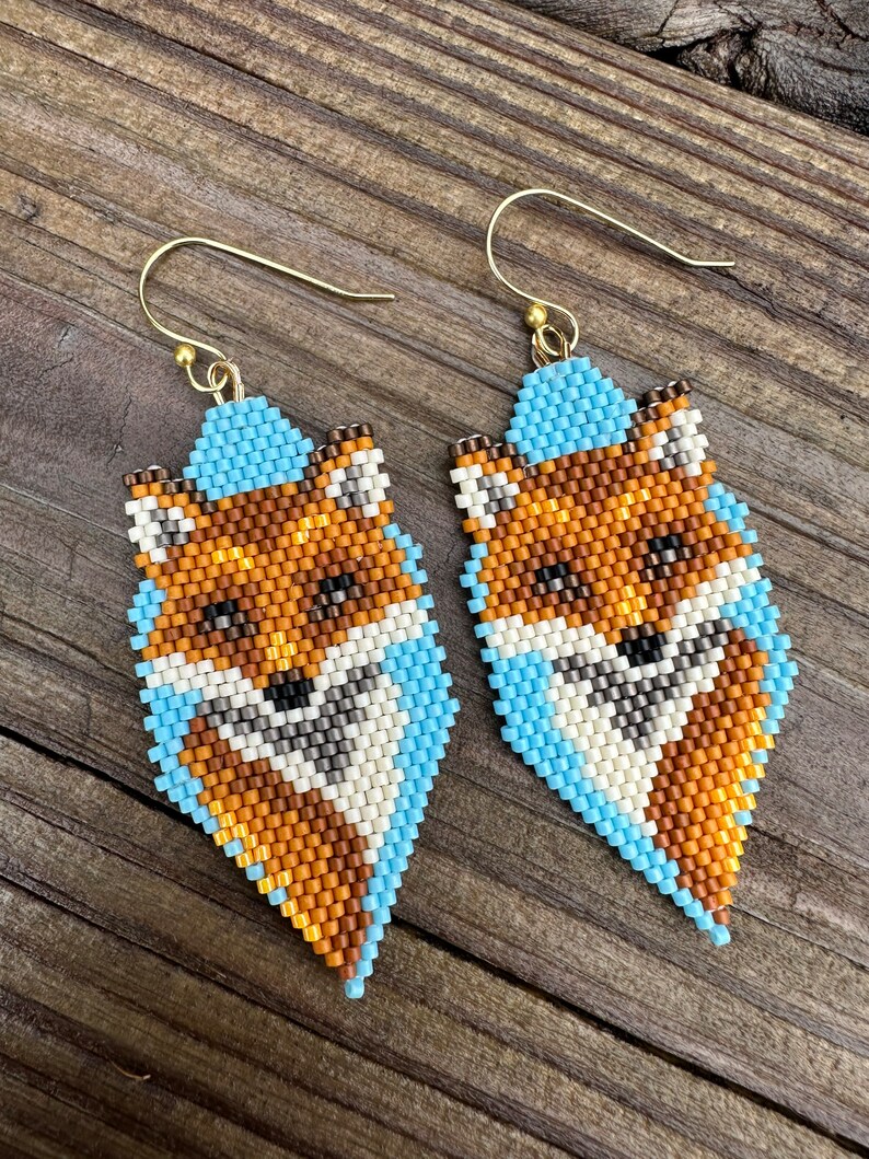 Puede incluir: Un par de pendientes de zorro con cuentas. Los pendientes presentan un dise&ntilde;o de zorro con cuentas naranjas, marrones, blancas y negras. El zorro est&aacute; delineado con cuentas de color azul claro y los pendientes tienen alambres dorados.