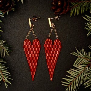 May include: A pair of red beaded heart-shaped earrings. The earrings are suspended from gold-colored posts and chains. The hearts are made of small, rectangular beads. The background is black, with pine needles and pine cones.