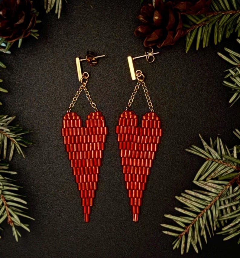 May include: A pair of red beaded heart-shaped earrings. The earrings are suspended from gold-colored posts and chains. The hearts are made of small, rectangular beads. The background is black, with pine needles and pine cones.