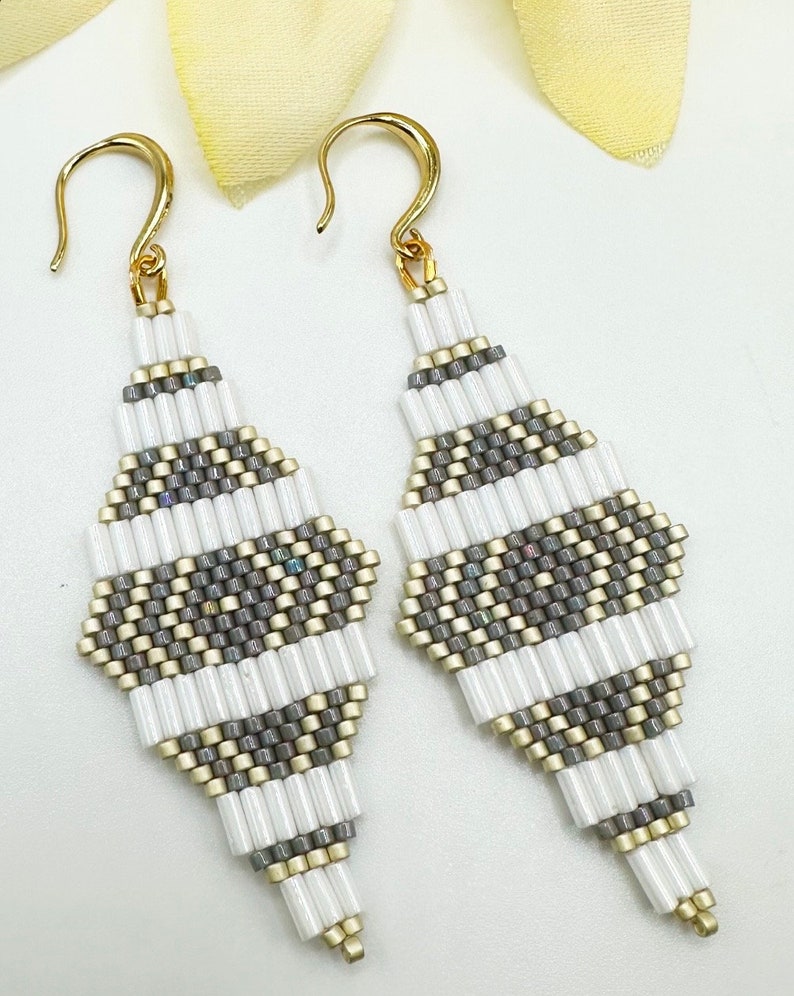 Pode incluir: Um par de brincos de contas brancas e cinzas com ganchos dourados. Os brincos t&ecirc;m a forma de um diamante e um padr&atilde;o geom&eacute;trico.