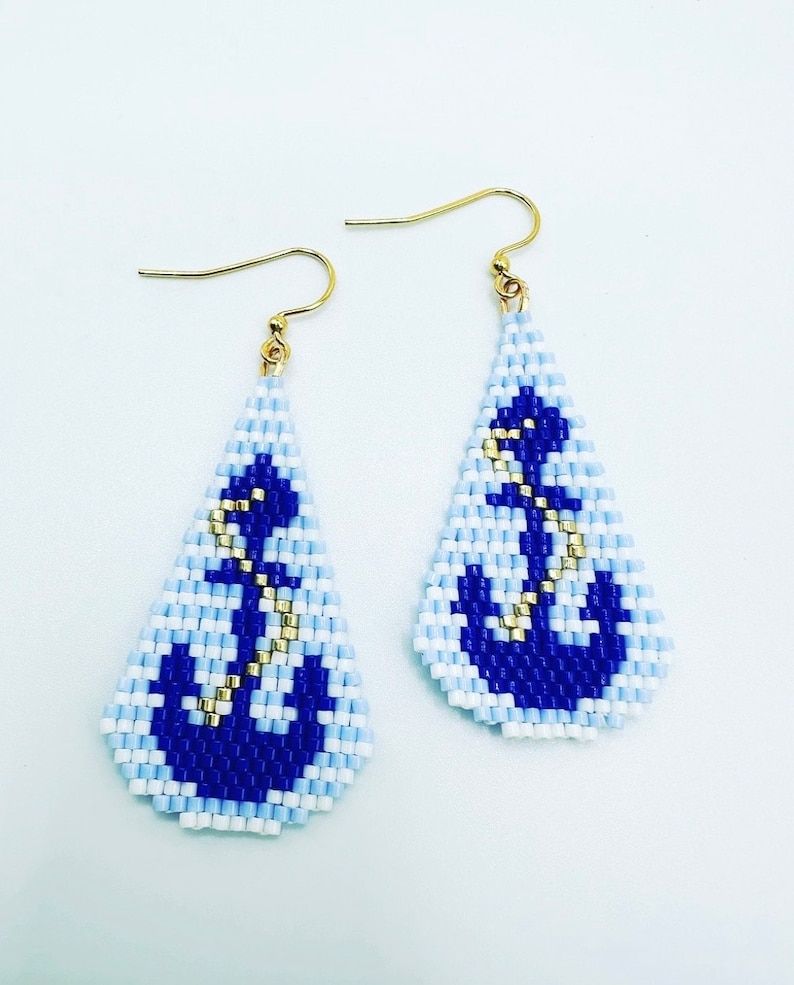 May include: A pair of teardrop earrings with a nautical theme. The earrings feature a beaded design with a blue anchor on a light blue and white background. Gold-coloured hooks complete the design.