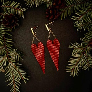 May include: A pair of red beaded heart-shaped earrings. The earrings are suspended from gold-colored bars and chains. The background is dark, with pine needles and pine cones framing the earrings.