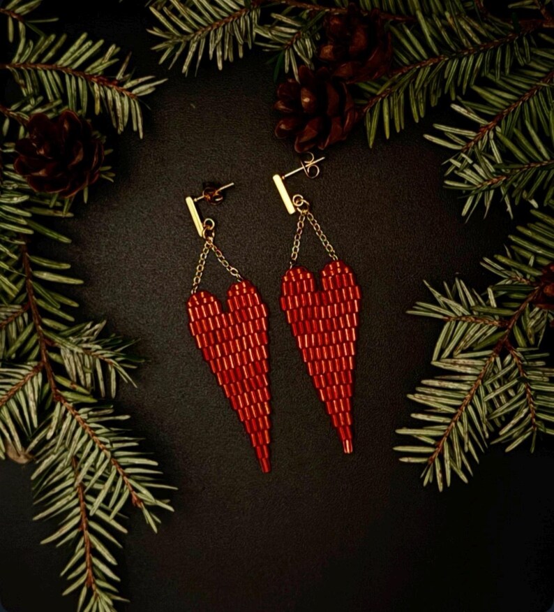 May include: A pair of red beaded heart-shaped earrings. The earrings are suspended from gold-colored bars and chains. The background is dark, with pine needles and pine cones framing the earrings.