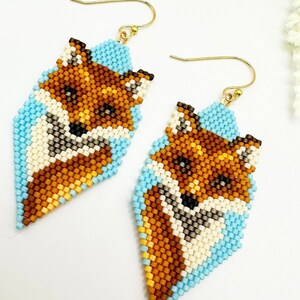 Puede incluir: Un par de pendientes de cuentas con dise&ntilde;o de zorro. Los pendientes presentan un dise&ntilde;o de zorro en tonos marr&oacute;n, crema y negro, sobre un fondo azul claro. Los pendientes tienen ganchos dorados.