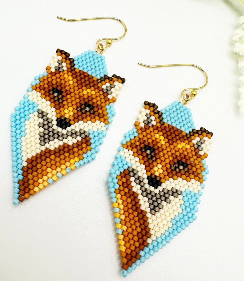 Puede incluir: Un par de pendientes de cuentas con dise&ntilde;o de zorro. Los pendientes presentan un dise&ntilde;o de zorro en tonos marr&oacute;n, crema y negro, sobre un fondo azul claro. Los pendientes tienen ganchos dorados.