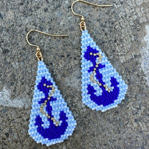 May include: Teardrop-shaped beaded earrings with a nautical anchor design. The earrings are light blue and dark blue, with gold-coloured accents. The earrings have gold-coloured ear wires.