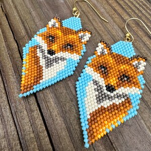 Puede incluir: Un par de pendientes de cuentas con dise&ntilde;o de zorro. Los pendientes presentan un dise&ntilde;o de zorro con cuentas naranjas, marrones, blancas y negras, sobre un fondo azul claro. Los pendientes tienen ganchos dorados.