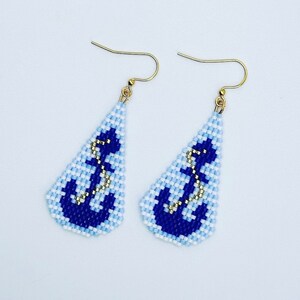 May include: A pair of beaded earrings with a nautical theme. Each earring has a light blue and white background, a dark blue anchor design, and gold-coloured hooks. The earrings are triangular in shape.