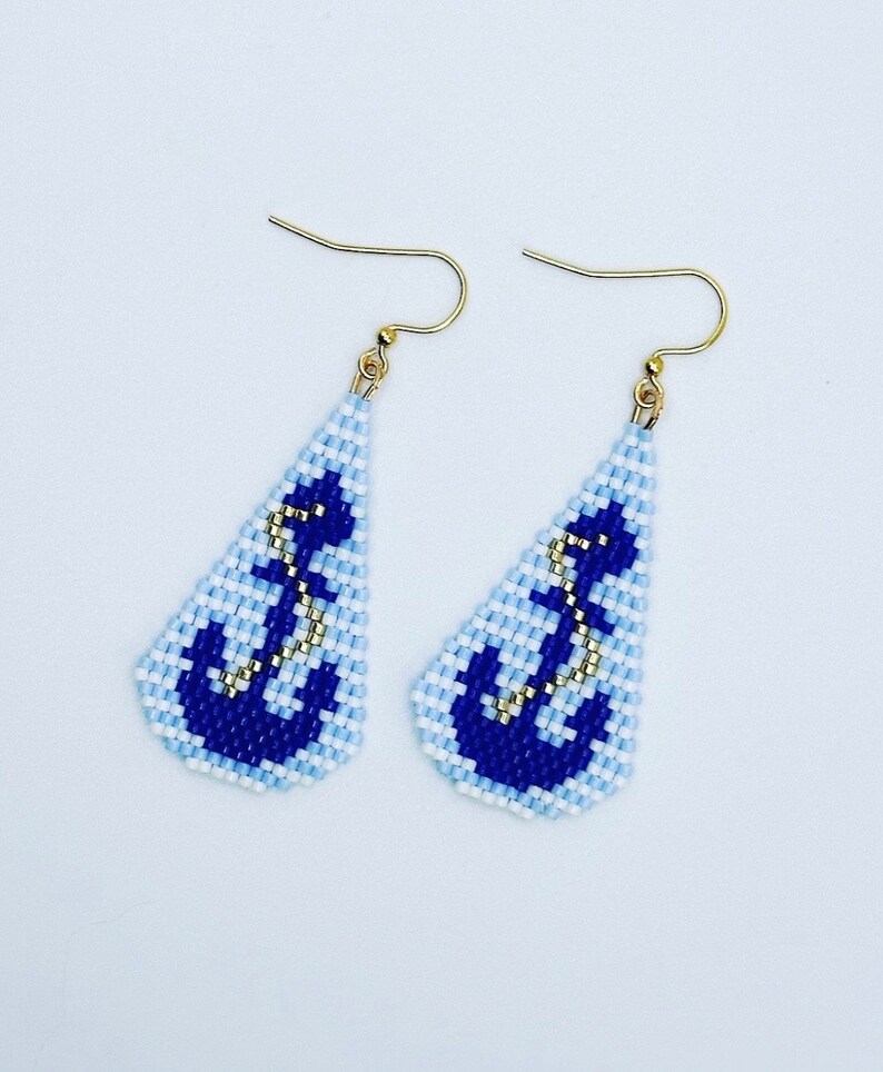 May include: A pair of beaded earrings with a nautical theme. Each earring has a light blue and white background, a dark blue anchor design, and gold-coloured hooks. The earrings are triangular in shape.