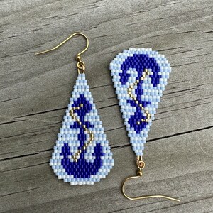 May include: A pair of beaded earrings with a nautical theme. The earrings feature a blue and white anchor design with gold-coloured accents. The earrings are teardrop-shaped and have gold-coloured ear wires.