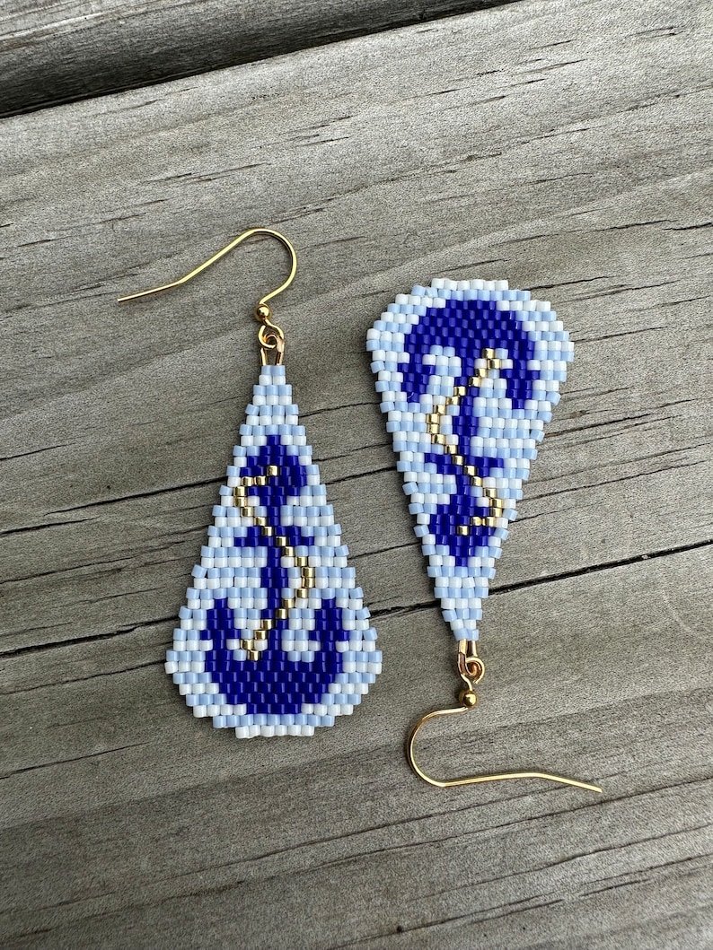 May include: A pair of beaded earrings with a nautical theme. The earrings feature a blue and white anchor design with gold-coloured accents. The earrings are teardrop-shaped and have gold-coloured ear wires.