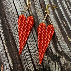 May include: A pair of red beaded heart earrings. The earrings are made of small, cylindrical beads and hang from gold-colored posts and chains. The heart shapes are long and tapered, with a gradient of red hues.