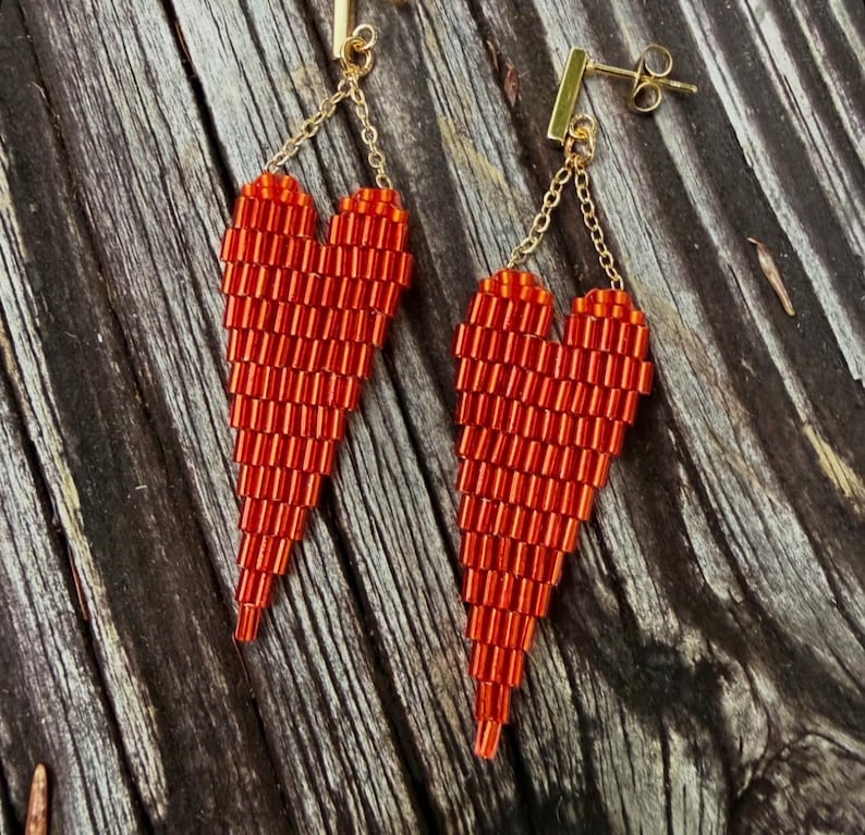May include: A pair of red beaded heart earrings. The earrings are made of small, cylindrical beads and hang from gold-colored posts and chains. The heart shapes are long and tapered, with a gradient of red hues.