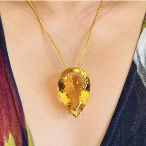 May include: A gold necklace featuring a large, teardrop-shaped, yellow gemstone pendant. The pendant is faceted, catching the light and displaying a brilliant, golden hue. The chain is a delicate gold color.