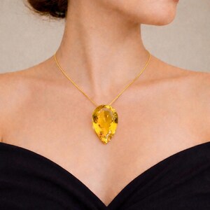 May include: A gold necklace featuring a large, pear-shaped, yellow gemstone pendant. The pendant is suspended from a delicate gold chain. The woman is wearing a black off-the-shoulder top.