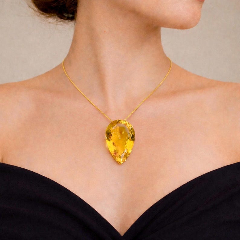 May include: A gold necklace featuring a large, pear-shaped, yellow gemstone pendant. The pendant is suspended from a delicate gold chain. The woman is wearing a black off-the-shoulder top.