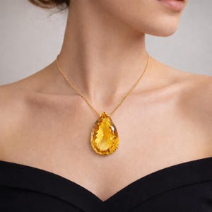 May include: A gold necklace featuring a large, pear-shaped, faceted yellow gemstone pendant. The pendant hangs from a delicate gold chain. The woman is wearing a black off-the-shoulder top.