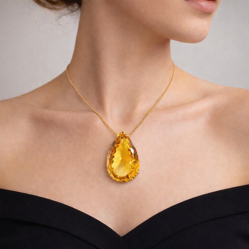May include: A gold necklace featuring a large, pear-shaped, faceted yellow gemstone pendant. The pendant hangs from a delicate gold chain. The woman is wearing a black off-the-shoulder top.