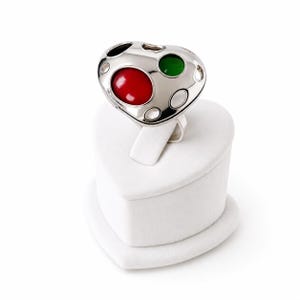 May include: A silver-toned ring featuring a heart-shaped face with a smooth, polished finish. The surface is accented with three circular, recessed inlays in vibrant red, deep green, and black, alongside several empty circular cutouts.