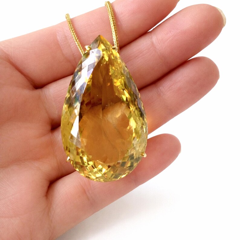 May include: A large, faceted, pear-shaped yellow gemstone pendant suspended from a delicate gold chain. The gemstone is a vibrant, translucent yellow with multiple facets that catch the light. The pendant is held in a hand against a white background.