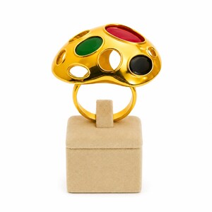 May include: A gold-tone ring featuring a wide, domed top with a series of circular cutouts. Three of these cutouts are filled with smooth, oval-shaped cabochon stones in red, green, and black, while the remaining cutouts are open and empty.