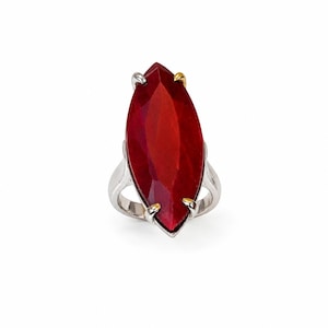 Può includere: Anello con una grande pietra rossa taglio marquise. L'anello ha una fascia argentata con dettagli dorati nella parte superiore e inferiore. La gemma è sfaccettata. Sfondo bianco.