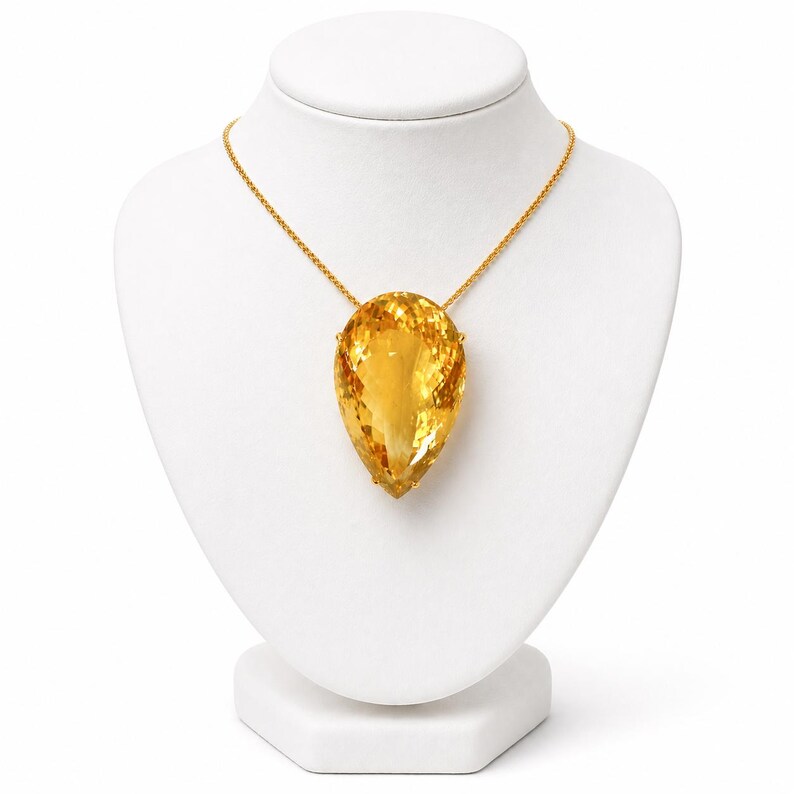 May include: A golden necklace featuring a large, pear-shaped citrine pendant. The pendant is a vibrant yellow-orange color and is suspended from a delicate gold chain. The necklace is displayed on a white jewelry bust.