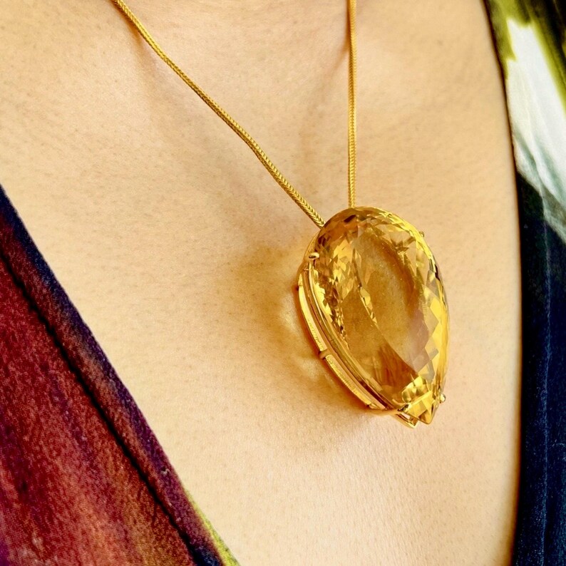 May include: A gold necklace featuring a large, faceted, teardrop-shaped pendant. The pendant is a warm, amber color, and the gold chain is delicate. The necklace is worn against a skin tone background, with a patterned fabric visible on the sides.