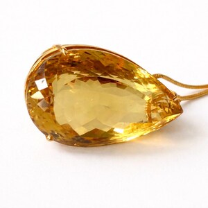 May include: A large, faceted, teardrop-shaped citrine gemstone pendant with a gold setting and chain. The gem is a vibrant yellow color, and the gold chain is delicate. The pendant is set against a white background.