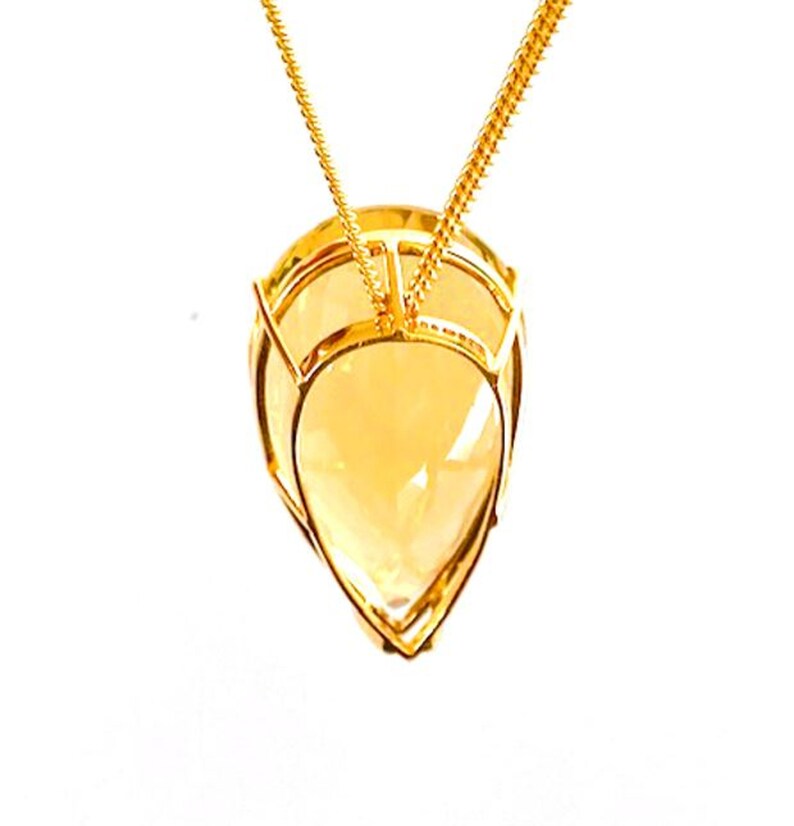 May include: A gold-colored pendant necklace featuring a large, teardrop-shaped, faceted, yellow gemstone. The gemstone is encased in a gold wire frame and hangs from a delicate gold chain. The pendant has a bright, polished finish.