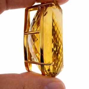 May include: A large, faceted, golden-yellow gemstone pendant held between fingers. The gemstone is encased in a gold-colored metal frame. The pendant has a geometric, rectangular shape with a diamond-cut pattern.