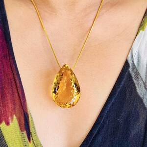 May include: A gold necklace featuring a large, faceted, teardrop-shaped citrine pendant. The pendant is a vibrant yellow-orange color and hangs from a delicate gold chain. The necklace is displayed against a patterned background.