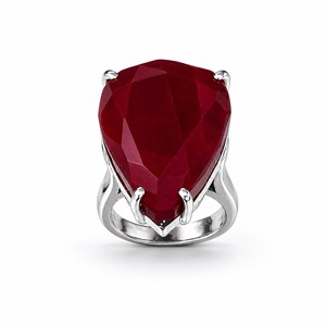 May include: A large, faceted, teardrop-shaped ruby gemstone ring. The deep red stone is set in a silver-colored metal band with four prongs securing the gem. The ring is displayed against a white background.