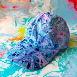 May include: A baseball cap with a vibrant, abstract pattern in shades of blue, purple, and pink. The cap features a curved brim and a classic six-panel design. The background has a similar swirling pattern.